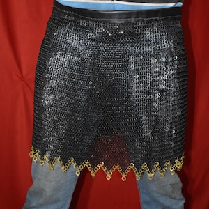 May include: A black chainmail skirt with a leather waistband and a decorative gold-colored trim along the bottom edge. The skirt is made of small, interlocking metal rings. The garment is shown against a red background.