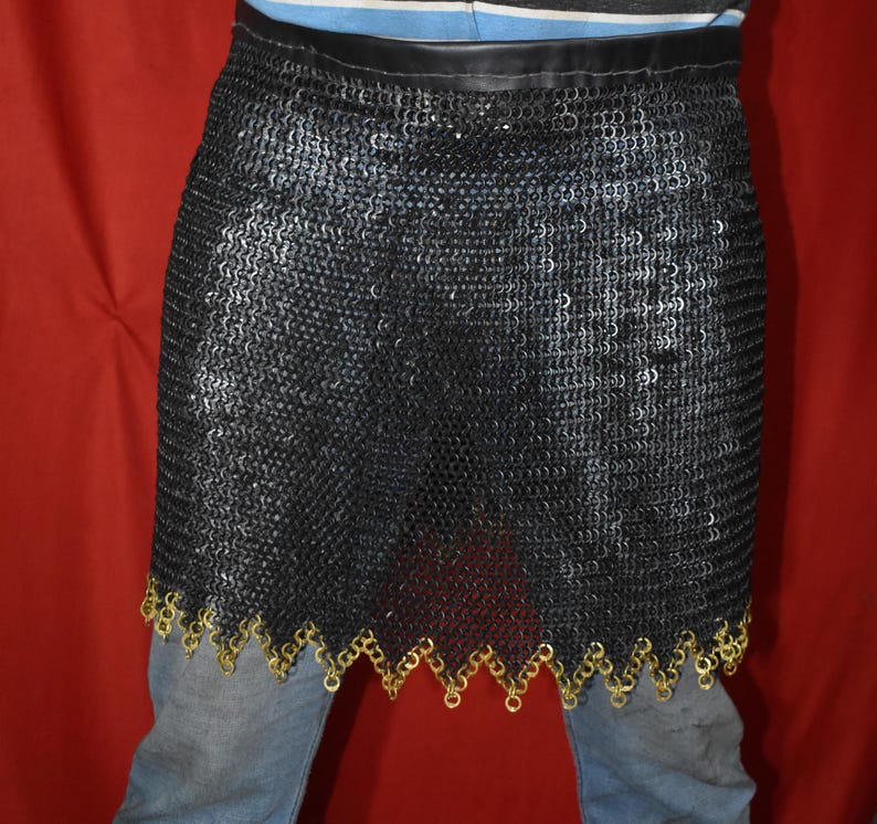 May include: A black chainmail skirt with a leather waistband and a decorative gold-colored trim along the bottom edge. The skirt is made of small, interlocking metal rings. The garment is shown against a red background.