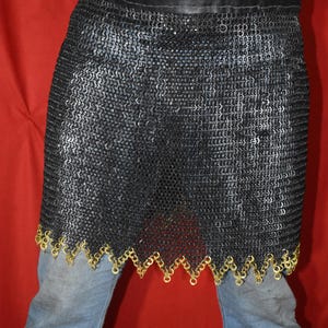 Puede incluir: Una falda de cota de malla gris oscuro con un cinturón de cuero negro. La falda tiene un ribete decorativo de color dorado a lo largo del borde inferior. La persona lleva vaqueros azules y una camisa a rayas azules y blancas.