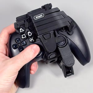 May include: Black gaming controller with a black Akaki attachment. The controller features a directional pad, action buttons, and analogue sticks. The Akaki attachment is designed to hold the controller.