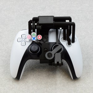 Puede incluir: Un mando de videojuegos blanco y negro con un accesorio negro impreso en 3D. El accesorio tiene cuatro brazos que se extienden desde la parte superior del mando. El mando tiene cuatro botones en la parte superior, cada uno con un s&iacute;mbolo de color diferente.