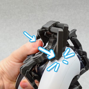 Puede incluir: Un accesorio de mando negro de pl&aacute;stico con una palanca que se puede mover para presionar los botones de un mando de videojuego blanco. El accesorio est&aacute; dise&ntilde;ado para ayudar a las personas con movilidad limitada de las manos a jugar a videojuegos.