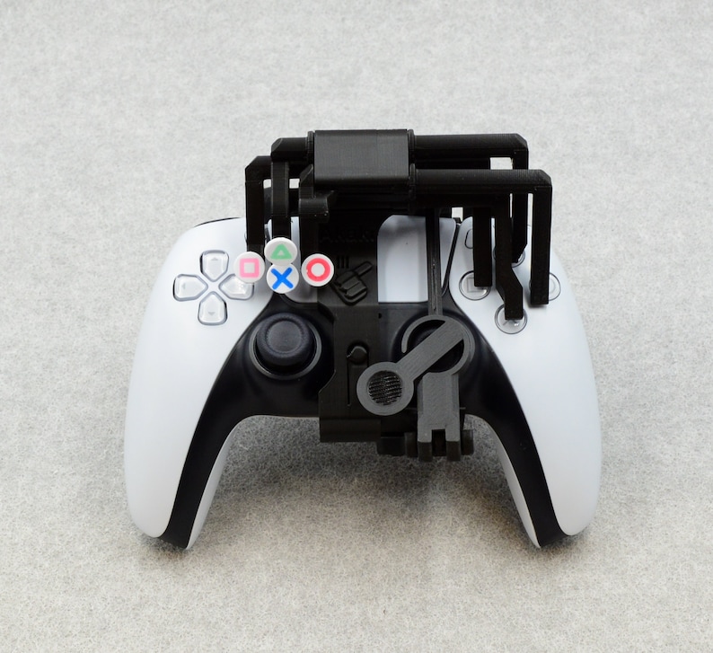 Puede incluir: Un mando de videojuegos blanco y negro con un accesorio negro impreso en 3D. El accesorio tiene cuatro brazos que se extienden desde la parte superior del mando. El mando tiene cuatro botones en la parte superior, cada uno con un s&iacute;mbolo de color diferente.