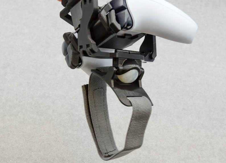 Puede incluir: Un soporte para auriculares VR impreso en 3D de color negro y gris con una correa gris. El soporte tiene una bola blanca en la parte inferior.