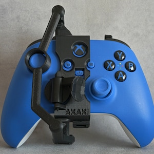 Może przedstawiać: Niebieski kontroler Xbox z czarnym mocowaniem. Kontroler ma kolorowe przyciski i joystick. Czarne mocowanie ma zakrzywione ramię i klips. Słowo "AKAKI" jest wydrukowane na czarnym mocowaniu.