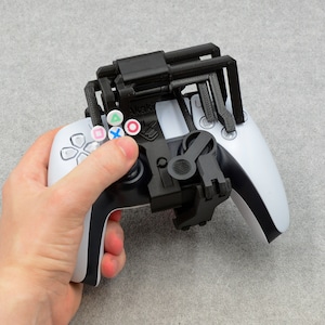 May include: A white PlayStation 5 DualSense controller with a black 3D printed accessory attached. The accessory has four prongs that extend from the top of the controller and a small lever on the right side. The controller has coloured buttons with the symbols: triangle, circle, cross, and square.