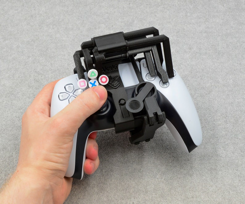 Puede incluir: Un mando de PlayStation 5 DualSense blanco con un accesorio impreso en 3D negro. El accesorio tiene cuatro puntas que se extienden desde la parte superior del mando y una peque&ntilde;a palanca en el lado derecho. El mando tiene botones de colores con los s&iacute;mbolos: tri&aacute;ngulo, c&iacute;rculo, cruz y cuadrado.