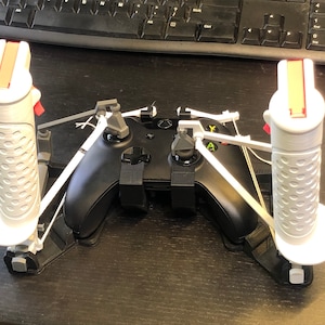May include: A black Xbox controller with two white 3D printed handles attached to it. The handles have red and orange accents. The controller is on a black surface.