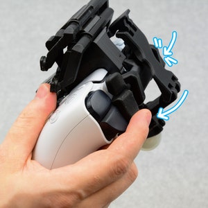 Puede incluir: Un accesorio de agarre para tel&eacute;fono de pl&aacute;stico negro para un mando de juego blanco. El agarre tiene una correa que se puede ajustar para adaptarse a diferentes tama&ntilde;os de mano. El agarre est&aacute; dise&ntilde;ado para fijarse en la parte posterior del mando, proporcionando un agarre m&aacute;s seguro.