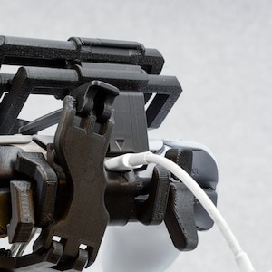 Puede incluir: Un soporte para tel&eacute;fono o mando impreso en 3D de color negro con un cable blanco conectado.