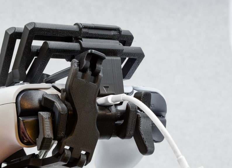Puede incluir: Un soporte para tel&eacute;fono o mando impreso en 3D de color negro con un cable blanco conectado.