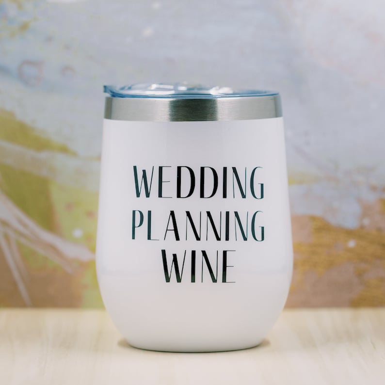 Puede incluir: Vaso de vino blanco sin tallo con borde plateado y tapa azul. El vaso tiene las palabras "WEDDING PLANNING WINE" impresas en negro. El fondo es una mezcla de blanco, dorado y azul claro.
