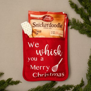 Puede incluir: Manopla de horno roja con el texto "We whisk you a Merry Christmas" y un dise&ntilde;o de batidor blanco. Un paquete de mezcla para galletas Snickerdoodle Betty Crocker y una esp&aacute;tula de silicona blanca est&aacute;n en el bolsillo de la manopla.
