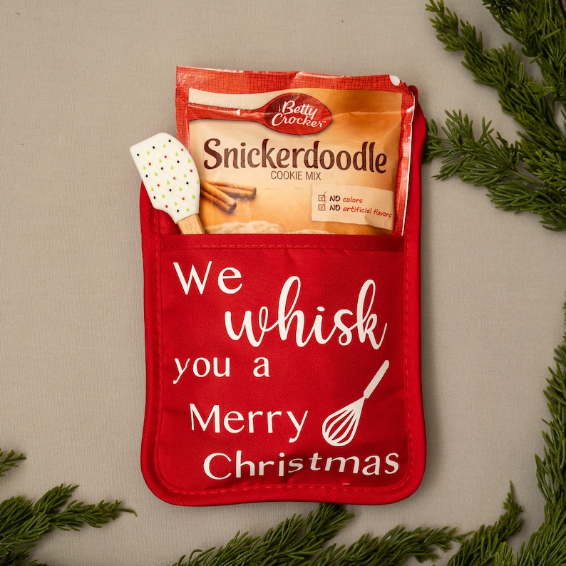 Puede incluir: Manopla de horno roja con el texto "We whisk you a Merry Christmas" y un dise&ntilde;o de batidor blanco. Un paquete de mezcla para galletas Snickerdoodle Betty Crocker y una esp&aacute;tula de silicona blanca est&aacute;n en el bolsillo de la manopla.