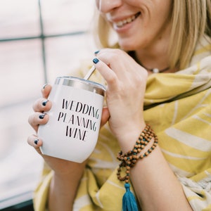 Puede incluir: Vaso de vino blanco de acero inoxidable con las palabras "WEDDING PLANNING WINE" en escritura negra. El vaso tiene una tapa plateada y una pajita. La persona lleva un chal con estampado amarillo y blanco y una pulsera de cuentas de madera.