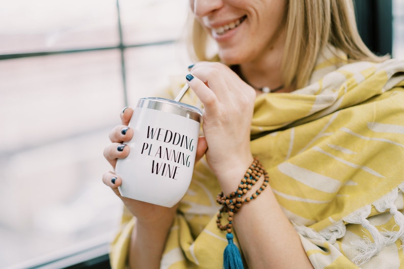 Puede incluir: Vaso de vino blanco de acero inoxidable con las palabras "WEDDING PLANNING WINE" en escritura negra. El vaso tiene una tapa plateada y una pajita. La persona lleva un chal con estampado amarillo y blanco y una pulsera de cuentas de madera.