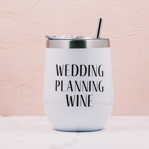 Puede incluir: Vaso de vino aislado blanco con borde y tapa plateados, y una pajita plateada. Las palabras "WEDDING PLANNING WINE" est&aacute;n impresas en negro en la parte delantera. El fondo es rosa claro.