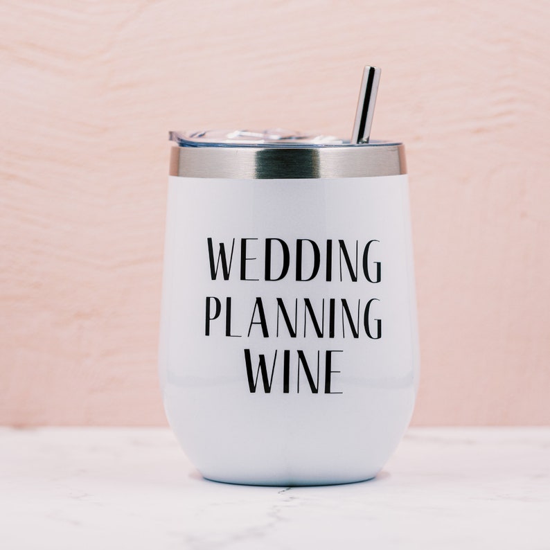 Puede incluir: Vaso de vino aislado blanco con borde y tapa plateados, y una pajita plateada. Las palabras "WEDDING PLANNING WINE" est&aacute;n impresas en negro en la parte delantera. El fondo es rosa claro.