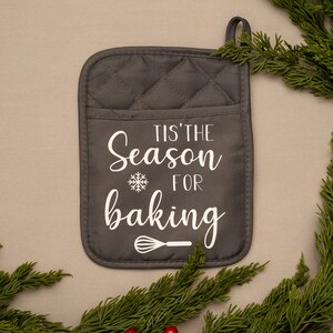 Puede incluir: Manopla de horno acolchada gris con el texto "Tis' the Season for baking" y un copo de nieve y un batidor.