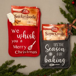 Puede incluir: Dos manoplas de horno rojas y grises con bolsillos. La manopla roja tiene el texto "We whisk you a Merry Christmas" y un gr&aacute;fico de batidor. La manopla gris tiene el texto "Tis' the Season for baking" y un gr&aacute;fico de copo de nieve. Ambas manoplas tienen un paquete de mezcla para galletas Betty Crocker en el bolsillo.