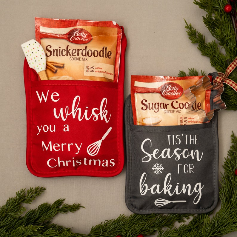 Puede incluir: Dos manoplas de horno rojas y grises con bolsillos. La manopla roja tiene el texto "We whisk you a Merry Christmas" y un gr&aacute;fico de batidor. La manopla gris tiene el texto "Tis' the Season for baking" y un gr&aacute;fico de copo de nieve. Ambas manoplas tienen un paquete de mezcla para galletas Betty Crocker en el bolsillo.