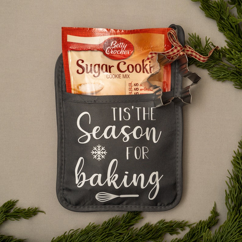 Puede incluir: Una manopla de horno gris con el texto "Tis' the Season for Baking" y un dise&ntilde;o de copo de nieve. Una cinta a cuadros roja y blanca est&aacute; atada alrededor de un paquete de mezcla para galletas de az&uacute;car Betty Crocker y un cortador de galletas plateado dentro de la manopla.