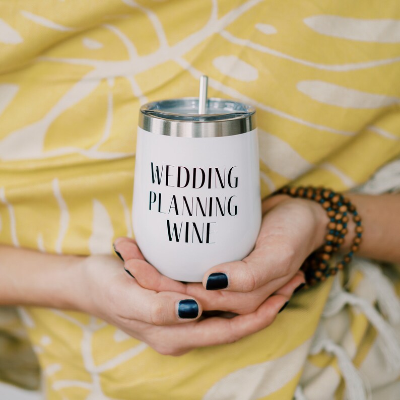 Puede incluir: Un vaso de vino blanco con borde y pajita plateados, con el texto "WEDDING PLANNING WINE" en negro. El vaso est&aacute; sostenido en manos con esmalte de u&ntilde;as azul oscuro, sobre un fondo floral amarillo.