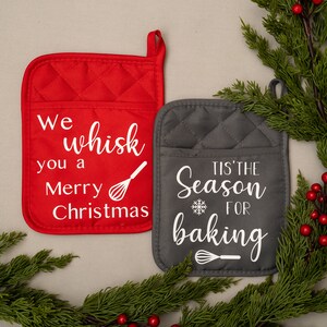 Puede incluir: Dos manoplas de horno rojas y grises con texto blanco. La manopla roja dice "We whisk you a Merry Christmas" con un gr&aacute;fico de batidor. La manopla gris dice "Tis' the Season for baking" con un copo de nieve y un gr&aacute;fico de batidor.