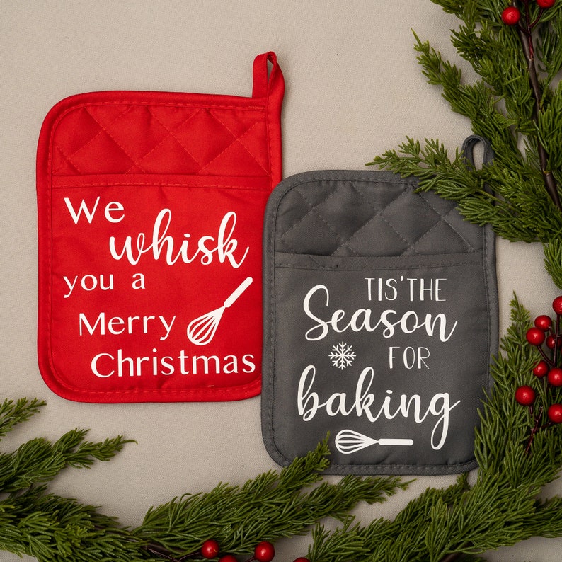 Puede incluir: Dos manoplas de horno rojas y grises con texto blanco. La manopla roja dice "We whisk you a Merry Christmas" con un gr&aacute;fico de batidor. La manopla gris dice "Tis' the Season for baking" con un copo de nieve y un gr&aacute;fico de batidor.