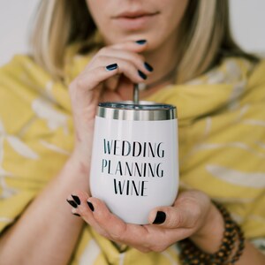 Puede incluir: Vaso de vino aislado blanco con el texto "WEDDING PLANNING WINE" en negro. El vaso tiene un borde plateado y lo sostiene una persona con esmalte de u&ntilde;as oscuro. El fondo incluye un chal amarillo.