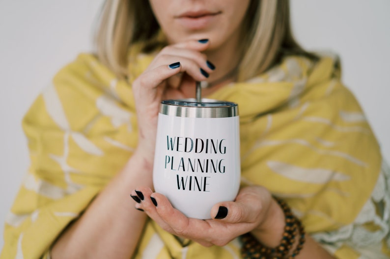 Puede incluir: Vaso de vino aislado blanco con el texto "WEDDING PLANNING WINE" en negro. El vaso tiene un borde plateado y lo sostiene una persona con esmalte de u&ntilde;as oscuro. El fondo incluye un chal amarillo.