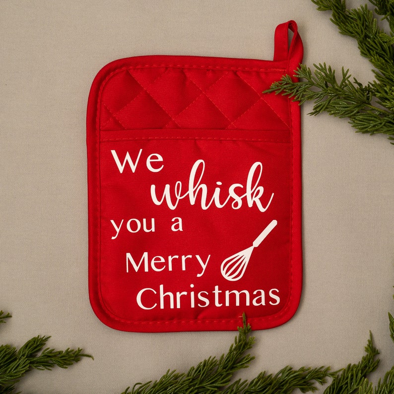 Puede incluir: Manopla de horno acolchada roja con un dise&ntilde;o blanco que dice "We whisk you a Merry Christmas" con un gr&aacute;fico de batidor blanco.
