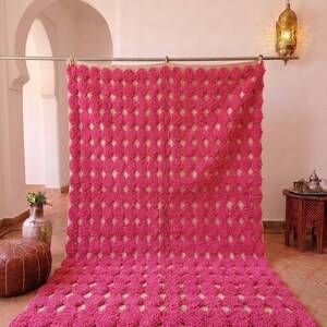 May include: A vibrant pink Moroccan rug with a geometric pattern of raised, circular tufts. The rug has a cream-colored base visible between the tufts and a fringe along the bottom edge. The rug is displayed on a tiled floor.