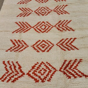 K&ouml;nnte beinhalten: Ein cremefarbener Teppich mit einem sich wiederholenden Muster aus roten geometrischen Formen. Das Design weist rauten- und pfeilartige Motive auf, die eine symmetrische und strukturierte Oberfl&auml;che schaffen. Der neutrale Hintergrund des Teppichs erg&auml;nzt die kr&auml;ftigen roten Akzente.