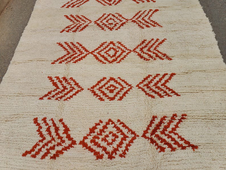 K&ouml;nnte beinhalten: Ein cremefarbener Teppich mit einem sich wiederholenden Muster aus roten geometrischen Formen. Das Design weist rauten- und pfeilartige Motive auf, die eine symmetrische und strukturierte Oberfl&auml;che schaffen. Der neutrale Hintergrund des Teppichs erg&auml;nzt die kr&auml;ftigen roten Akzente.