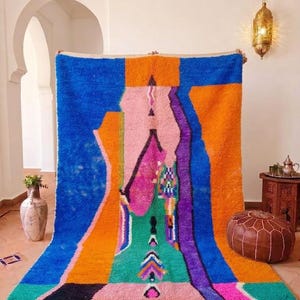 May include: A vibrant, abstract Moroccan rug with a bold color palette. The rug features a central design in shades of pink, green, and purple, framed by orange and blue sections. The rug has a fringed edge and is displayed indoors.