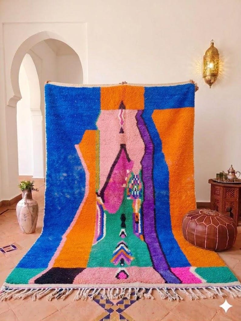May include: A vibrant, abstract Moroccan rug with a bold color palette. The rug features a central design in shades of pink, green, and purple, framed by orange and blue sections. The rug has a fringed edge and is displayed indoors.