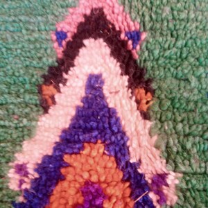 May include: Close-up of a section of a hand-woven rug. The rug features a geometric design with a green background and a central pattern in shades of pink, black, white, blue, orange, and purple. The texture appears soft and plush.