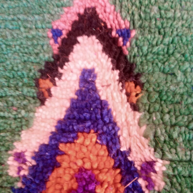 May include: Close-up of a section of a hand-woven rug. The rug features a geometric design with a green background and a central pattern in shades of pink, black, white, blue, orange, and purple. The texture appears soft and plush.