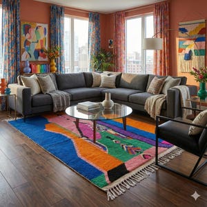 May include: A modern living room featuring a large gray sectional sofa, a round glass coffee table, and a vibrant, colorful area rug. The rug has a bold abstract design with blue, orange, green, and pink hues. The room is decorated with colorful curtains and artwork.