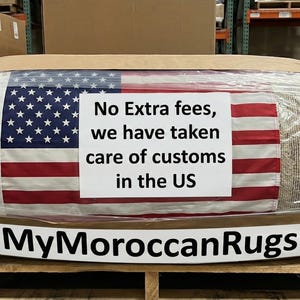 K&ouml;nnte beinhalten: Ein gerollter Teppich, verpackt in braunem Papier und durchsichtigem Kunststoff, mit Klebeband befestigt. Die amerikanische Flagge ist auf dem Teppich zu sehen, mit einem Schild mit der Aufschrift "No Extra fees, we have taken care of customs in the US." Der Markenname "MyMoroccanRugs" wird angezeigt.