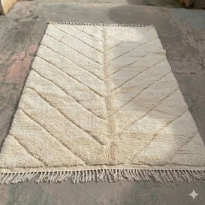Op de afbeelding: Een rechthoekig gebroken wit vloerkleed met een gestructureerd visgraatpatroon over het oppervlak. Het kleed is gemaakt met een dikke, hoogpolige weving en is afgewerkt met lange, geknoopte franjes aan zowel de boven- als onderrand.