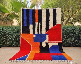 Puede incluir: Una alfombra rectangular de pelo largo con un diseño geométrico audaz. El patrón incorpora bloques y rayas en rojo vibrante, naranja brillante, azul intenso, negro y blanco crema, rematada con un borde de flecos anudados.