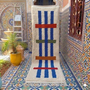 May include: A rectangular off-white area rug featuring three vertical blue stripes intersected by four horizontal bars in red, green, orange, and red. The rug has a thick, textured pile and a fringed edge at the bottom, displaying a geometric pattern.