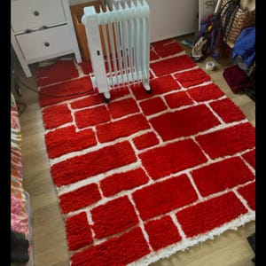 May include: A red brick-patterned rug with white grout lines. The rug is rectangular and has a shaggy texture. A white electric heater is placed on the rug. The rug is on a wooden floor.