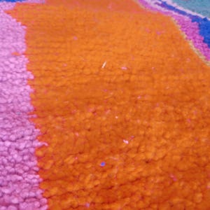May include: Close-up of a textured rug with vibrant colors. The rug features a large orange section, a pink section, and hints of blue and purple. The fibers are tightly woven, creating a plush appearance.