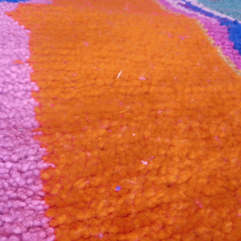 May include: Close-up of a textured rug with vibrant colors. The rug features a large orange section, a pink section, and hints of blue and purple. The fibers are tightly woven, creating a plush appearance.