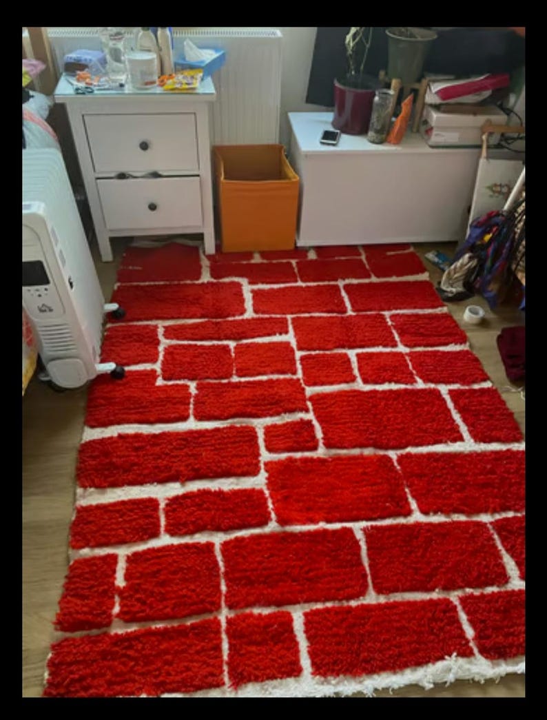 May include: A rectangular rug with a brick pattern. The rug features red rectangular shapes separated by white spaces, creating a brick wall design. The rug is placed on a wooden floor.