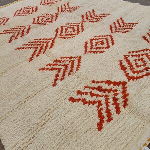K&ouml;nnte beinhalten: Cremefarbener Teppich mit einem sich wiederholenden Muster aus roten geometrischen Formen. Der Teppich hat eine strukturierte Oberfl&auml;che und einen gelben Rand mit wei&szlig;en Quasten. Das Design zeigt Diamant- und pfeilartige Motive.