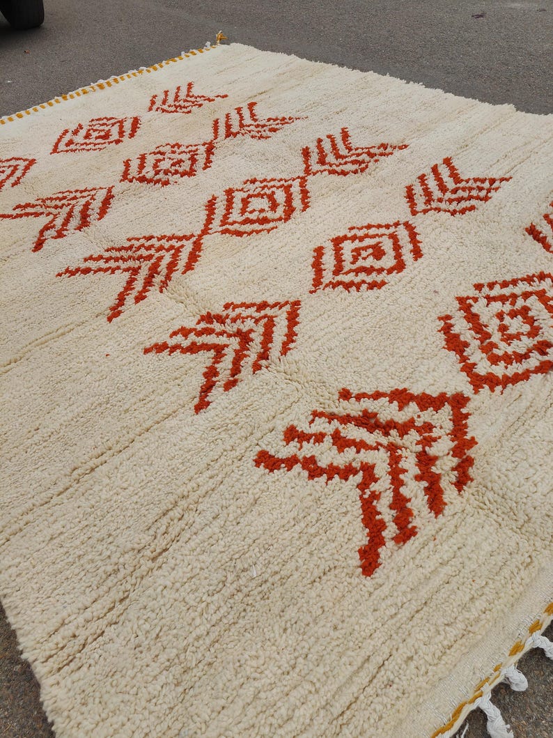K&ouml;nnte beinhalten: Cremefarbener Teppich mit einem sich wiederholenden Muster aus roten geometrischen Formen. Der Teppich hat eine strukturierte Oberfl&auml;che und einen gelben Rand mit wei&szlig;en Quasten. Das Design zeigt Diamant- und pfeilartige Motive.